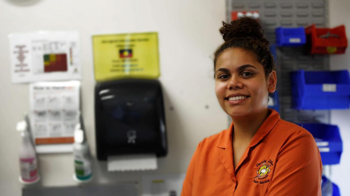Danila Dilba health worker sits in clinic smiling for photo. She is wearing an orange Danila Dilba work shirt.